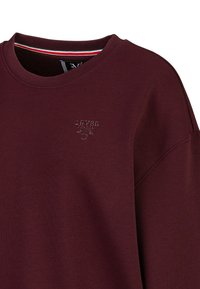 Bordeauxroter Sweatshirt mit Rundhalsausschnitt, verziert mit gesticktem Logo und gestreiftem Futter am Kragen in Rot, Weiß und Grün.