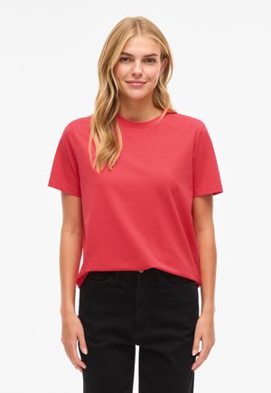 Blonde woman with loose hair wearing a plain red short-sleeve T-shirt and black pants, standing against a white background.