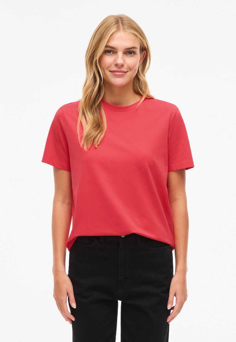 Vaaleatukkainen nainen, jolla on auki olevat hiukset, pukeutuneena yksiväriseen punaiseen lyhythihaiseen T-paitaan ja mustiin housuihin, seisomassa valkoisen taustan edessä.