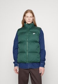 Grøn puffy vest med høj krave og lynlås foran, med sidelommer og et lille hvidt Nike-logo på venstre bryst. Båret over en blå denimskjorte.