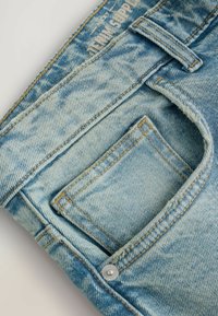 Világoskék farmernadrág, amelyen elhalványodás látható, elülső zsebbel és díszvarrással. Fémgomb található a derekán.