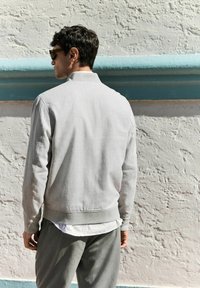 Chaqueta bomber gris con puños y dobladillo acanalados, con una tela texturizada. Llevada sobre una camisa blanca, combinada con pantalones grises.