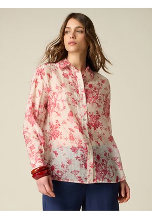 Frau mit welligem Haar, die eine transparente weiße Bluse mit rotem Blumenmuster und marineblaue Hose trägt, ergänzt mit roten Armreifen am linken Handgelenk.