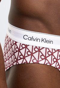 Męskie slipy Calvin Klein z białym paskiem w pasie, ozdobione czerwonym geometrycznym wzorem na różowym tle. Miękki, rozciągliwy materiał.