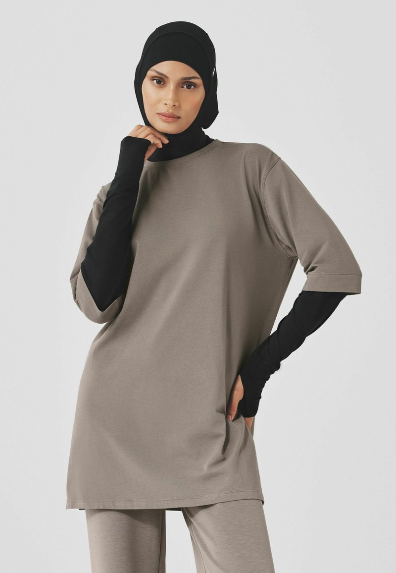 Oversized, korte mouwen top in taupe katoenmix met zijsplitsen. Draagt over een zwarte langemouwen basislaag, wat de gelaagde look benadrukt.