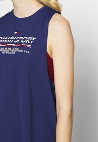 Top atletico senza maniche di colore blu navy con superficie texturizzata, caratterizzato da un logo del marchio bianco e rosso sul petto, indossato sopra una maglietta interna color bordeaux.