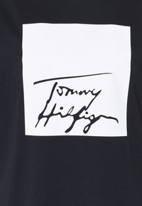 Camiseta negra con un parche rectangular blanco que presenta una firma estilizada en negro. La tela es suave; el diseño enfatiza el contraste.
