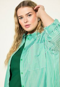 Camisa de botones a rayas verdes y blancas con un bolsillo en el pecho, superpuesta sobre una camiseta verde sólida. Tejido suave con un corte relajado.