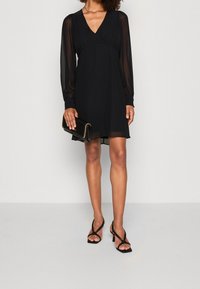 Schwarzes Kleid mit langen transparenten Ärmeln, V-Ausschnitt, tailliertem Bund und fließendem Rock. Kombiniert mit einer schwarzen Clutch und schwarzen Riemchenabsätzen.