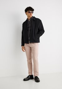 Νέος άνδρας με μαύρα γυαλιά ηλίου, μαύρο μπουφάν, σκούρο πουκάμισο, μπεζ παντελόνι και μαύρα παπούτσια, στέκεται μπροστά σε λευκό φόντο.