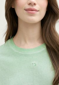 T-shirt vert clair avec un col rond, texture douce et logo brodé discret sur la poitrine. Le tissu semble légèrement décoloré.