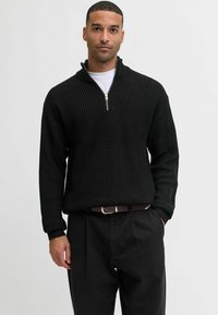 Suéter de punto negro con textura acanalada y cuello con media cremallera. Combinado con pantalones negros y un cinturón marrón, con un ajuste relajado.