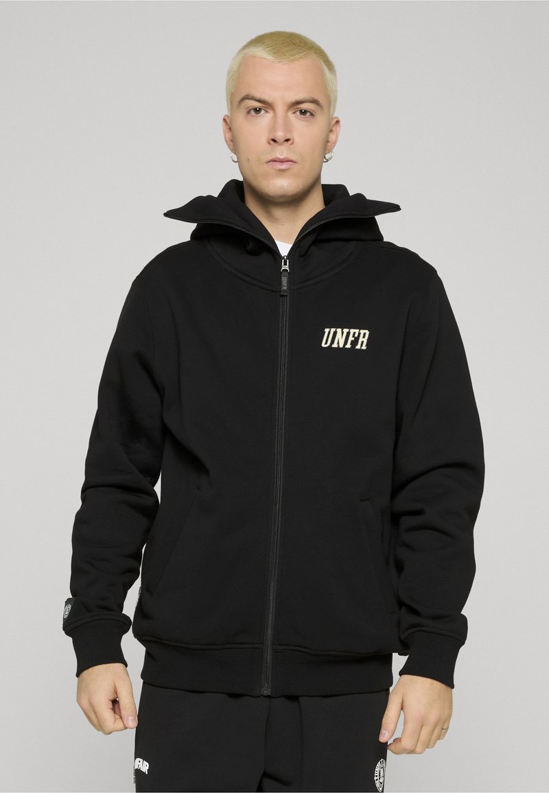 Jeune personne aux cheveux blonds courts portant un sweat à capuche noir zippé avec le texte « UNFR » sur la poitrine et un pantalon noir assorti.