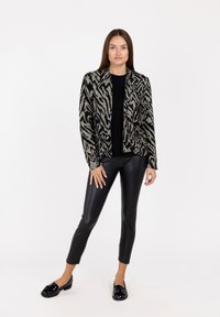 Cardigan a strisce zebrate nero e crema con un design in maglia testurizzata, caratterizzato da una parte anteriore senza colletto, indossato sopra un top nero e abbinato a pantaloni neri.