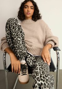 Pulover bej tricotat, cu gât elastic, asortat cu pantaloni cu imprimeu leopard alb-negru. Modelul poartă adidași bej deschis cu șireturi negre.