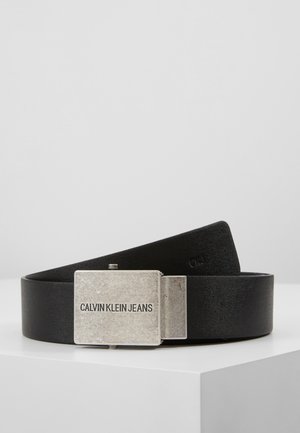 Czarny materiałowy pasek z prostokątną srebrną metalową klamrą z wygrawerowanym napisem "CALVIN KLEIN JEANS", przedstawiony na białej powierzchni.