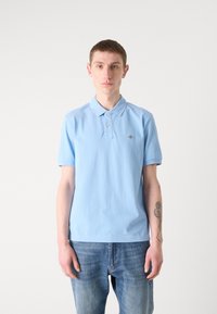 Lyseblå polo-skjorte laget av bomull, med krage og to-knapps placket, sammen med falmede blå jeans. Diskret logo på brystet.
