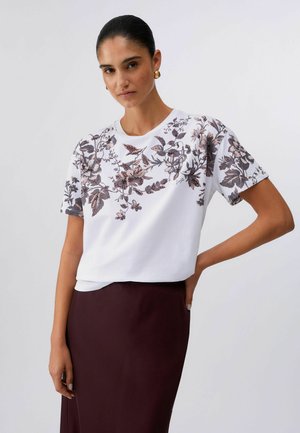 Donna con capelli scuri che indossa una maglia bianca a maniche corte con stampa floreale marrone e gonna marrone scuro, in piedi contro uno sfondo chiaro e uniforme.