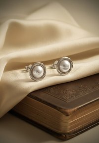 Runda silverörhängen med en slät vit pärla i mitten, vilande på en krämfärgad tyg över ett strukturerat brunt bokomslag.