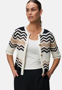 Strikket cardigan med et chevronmønster i beige, sort og hvid. Har korte ærmer, guldknapper og et struktureret design.