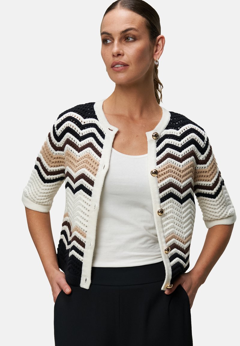 Strikket cardigan med et chevronmønster i beige, sort og hvid. Har korte ærmer, guldknapper og et struktureret design.