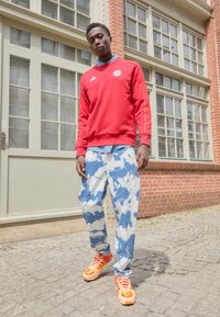 Rød hettegenser med hvite Adidas-striper, blå tie-dye bukser og livlige oransje joggesko. Modellen står på brosteinsbelagt område nær en murvegg.