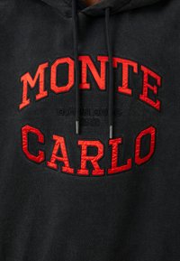 Must särg punase tikandiga tekstiga "MONTE CARLO" ja väiksema musta tekstiga "BJÖRN BORG 1956" kaela all, millel on tõmblukud.