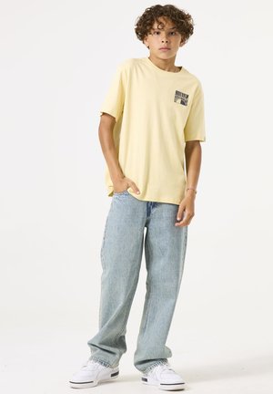 Gelbes T-Shirt mit kleiner schwarzer Grafik auf der Brusttasche, kombiniert mit hellblauen, weiten Jeans und weißen Sneakers. Einfaches Design mit lässiger Passform.