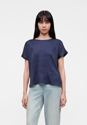 Jeune femme aux cheveux noirs raides, portant un chemisier ample bleu foncé à manches courtes et un jean bleu clair, debout devant un fond uni.