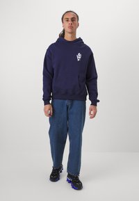 Marineblå hettegenser med frontlomme og grafisk trykk på brystet, kombinert med løse blå denimjeans og svarte sneakers med blå detaljer.