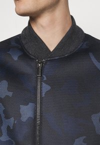 Närbild av en person som bär en svart nätjacka med blå kamouflagemönster, med hög krage och dragkedja framtill.