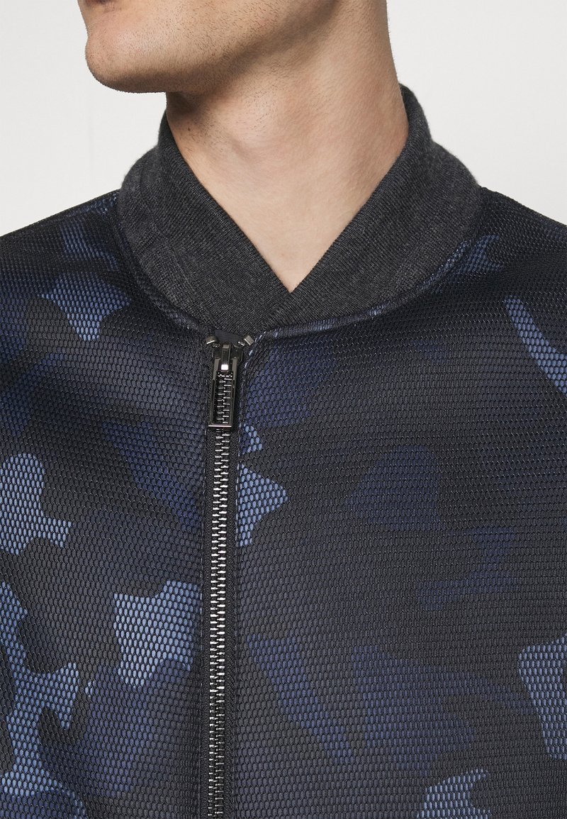 Närbild av en person som bär en svart nätjacka med blå kamouflagemönster, med hög krage och dragkedja framtill.