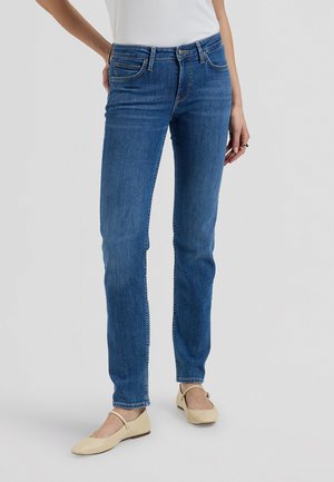 Kobieta ubrana w dopasowane niebieskie dżinsy, biały top i beżowe płaskie buty, stojąca z jedną ręką na biodrze na jednolitym tle.