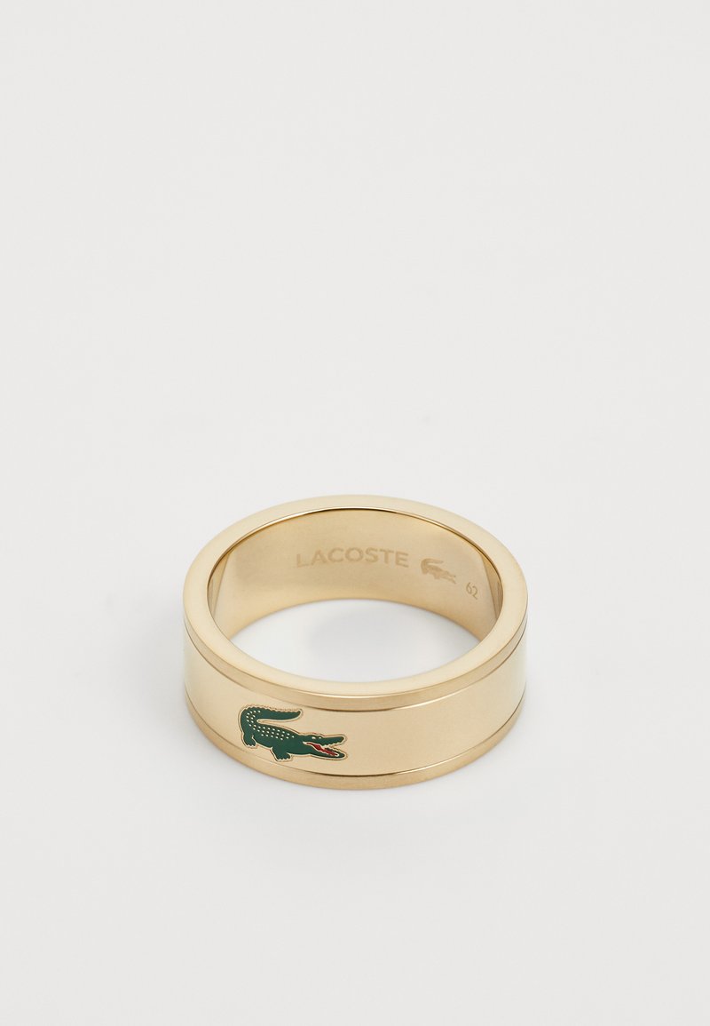 Auksinės spalvos žiedas su lygiu, blizgiu paviršiumi, kuriame yra žalias krokodilo logotipas ir viduje išgraviruotas prekės ženklas "LACOSTE". Dydis 62.