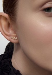 Kolczyk w kształcie liścia pokryty złotem z misternymi detalami, umieszczony na lekko widocznym uchu. Gładka faktura, mały i delikatny design.