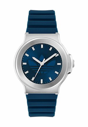 Blå silikonestrap ure med et sølvfarvet rustfrit stålhus, der har en mørkeblå urskive, sølvfarvede timemarkeringer og et logodesign.