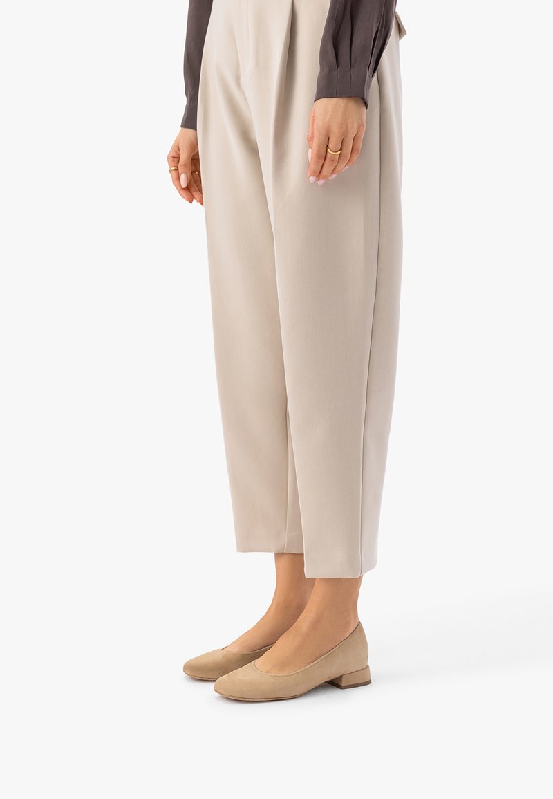 Osoba ubrana w beżowe spodnie 7/8 z plisami oraz beżowe zamszowe buty na niskim obcasie, stojąca bokiem z rękami swobodnie opuszczonymi wzdłuż ciała.