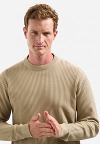 Beige geribbelde gebreide trui met een ronde halslijn en lange mouwen, met een textuurstof en aansluitende manchetten. Model houdt de handen voor zich gekleed.