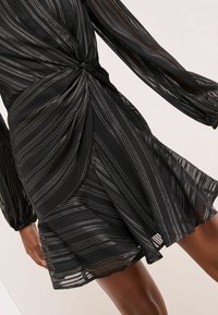 Černé průhledné šaty se stříbrnými vertikálními pruhy, zkroucený uzel v pase a dlouhé nafouklé rukávy na osobě s tmavou pletí.