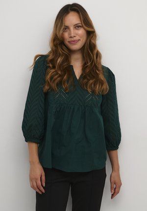 Blusa verde scuro con maniche a tre quarti, caratterizzata da un colletto a V e un intricato motivo ad occhio di bue. Tessuto morbido, vestibilità ampia e vita arricciata.
