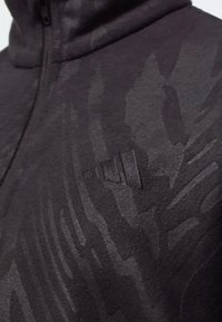 Hoodie zippé noir avec un motif camouflage texturé, doté d'un logo en relief sur la poitrine gauche. Matière douce avec une finition lisse.