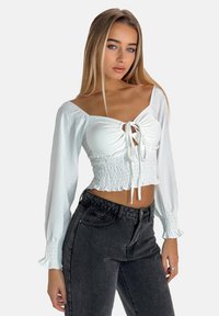 Top court blanc avec un design froncé, un devant noué et de longues manches bouffantes. Associe à un jean taille haute gris foncé pour le contraste.