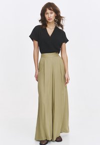 Czarna bluzka z dekoltem w serek i krótkimi rękawami, zestawiona z kieszonkowymi spodniami o wysokim stanie w kolorze oliwkowym i szerokimi nogawkami. Modelka stoi w neutralnym otoczeniu.