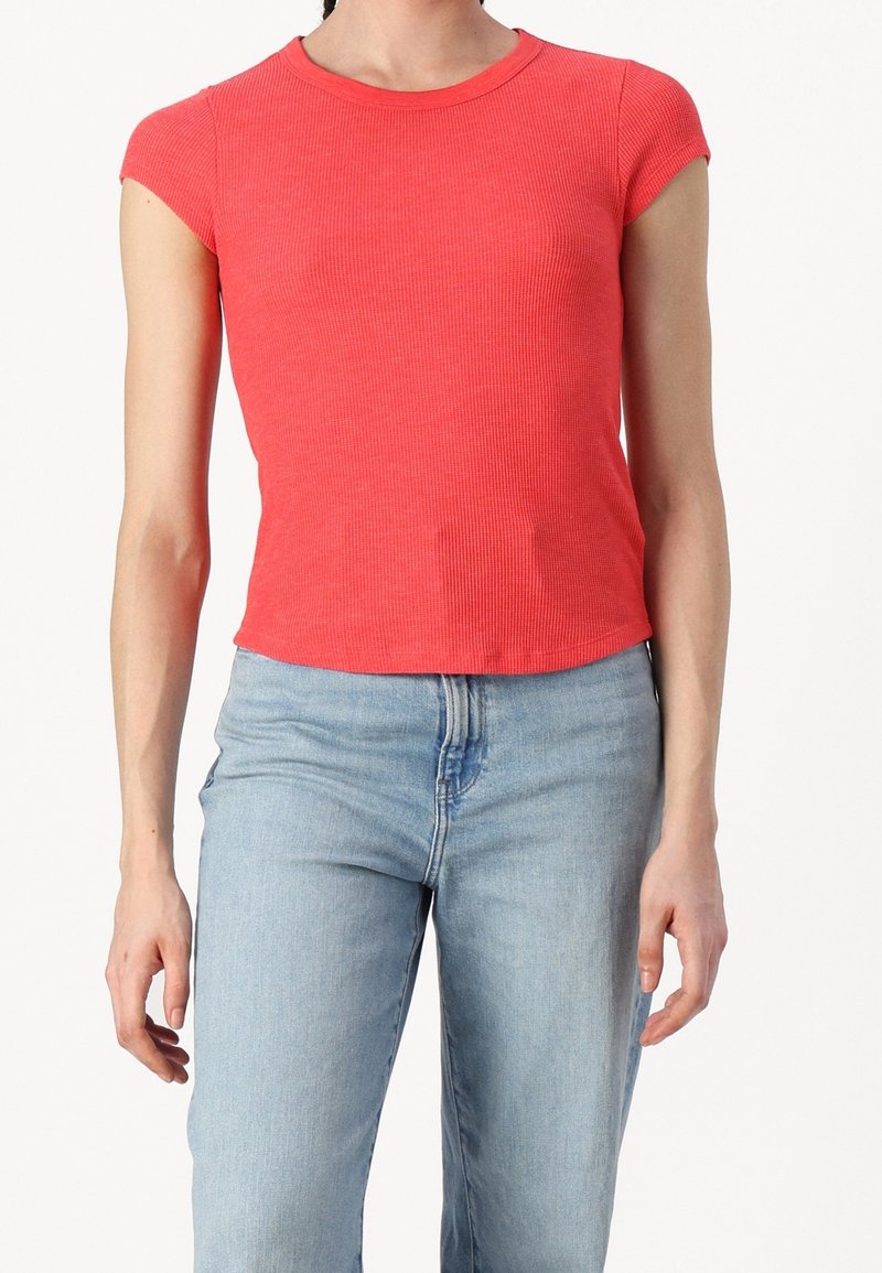 Personne portant un top rouge côtelé ajusté à manches courtes, associé à un jean bleu clair taille haute, sur un fond blanc uni.