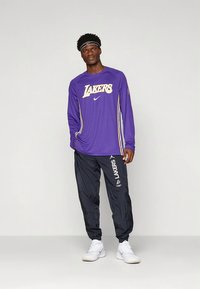 Violetti pitkähihaispaita, jossa on "LAKERS"-grafiikka ja valkoinen Nike-logo, yhdistettynä mustiin kevyisiin housuihin, joissa on valkoinen "LAKERS"-teksti.