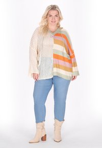 Poncho in maglia con scollo a V, caratterizzato da strisce multicolori in arancione, verde e crema, abbinato a jeans azzurri e stivaletti beige.