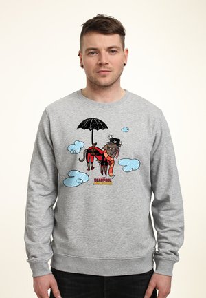Mężczyzna w szarej bluzie z kapturem z kreskówkowym psem przebranym za Deadpoola trzyma parasol, otoczony niebieskimi chmurami, stoi na jednolitym tle.