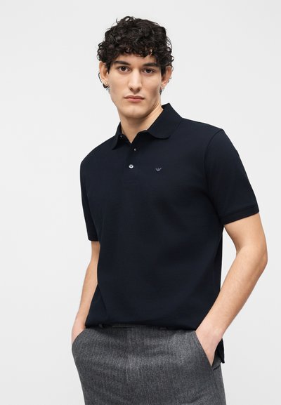 Νέος άνδρας με σγουρά σκούρα μαλλιά, ντυμένος με μαύρο πόλο πουκάμισο και γκρι παντελόνι, στέκεται με τα χέρια στις τσέπες μπροστά σε απλό φόντο.