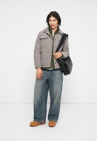 Veste grise matelassée avec poches avant, superposée à une chemise verte. Jean en denim large et chaussures beiges. Tient un sac à bandoulière noir.