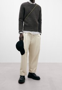 Suéter marrón de punto acanalado, camisa blanca de cuello con rayas, pantalones de pierna ancha beige, zapatos negros y sosteniendo una gorra negra. Diseño minimalista.
